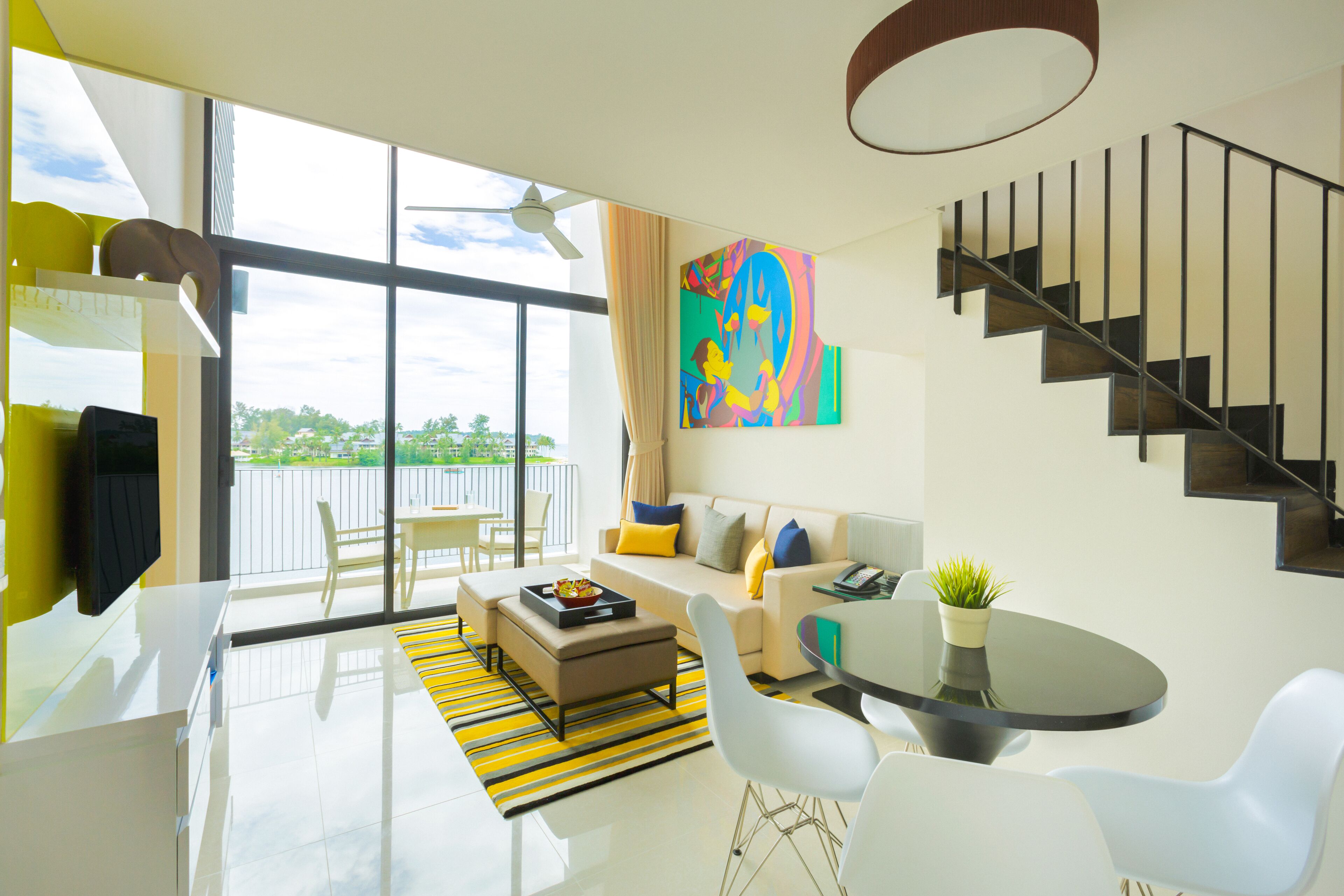 Una sala moderna con sofá, mesa de centro y sillas, un comedor con mesa y sillas, una escalera y ventanas grandes con vistas.