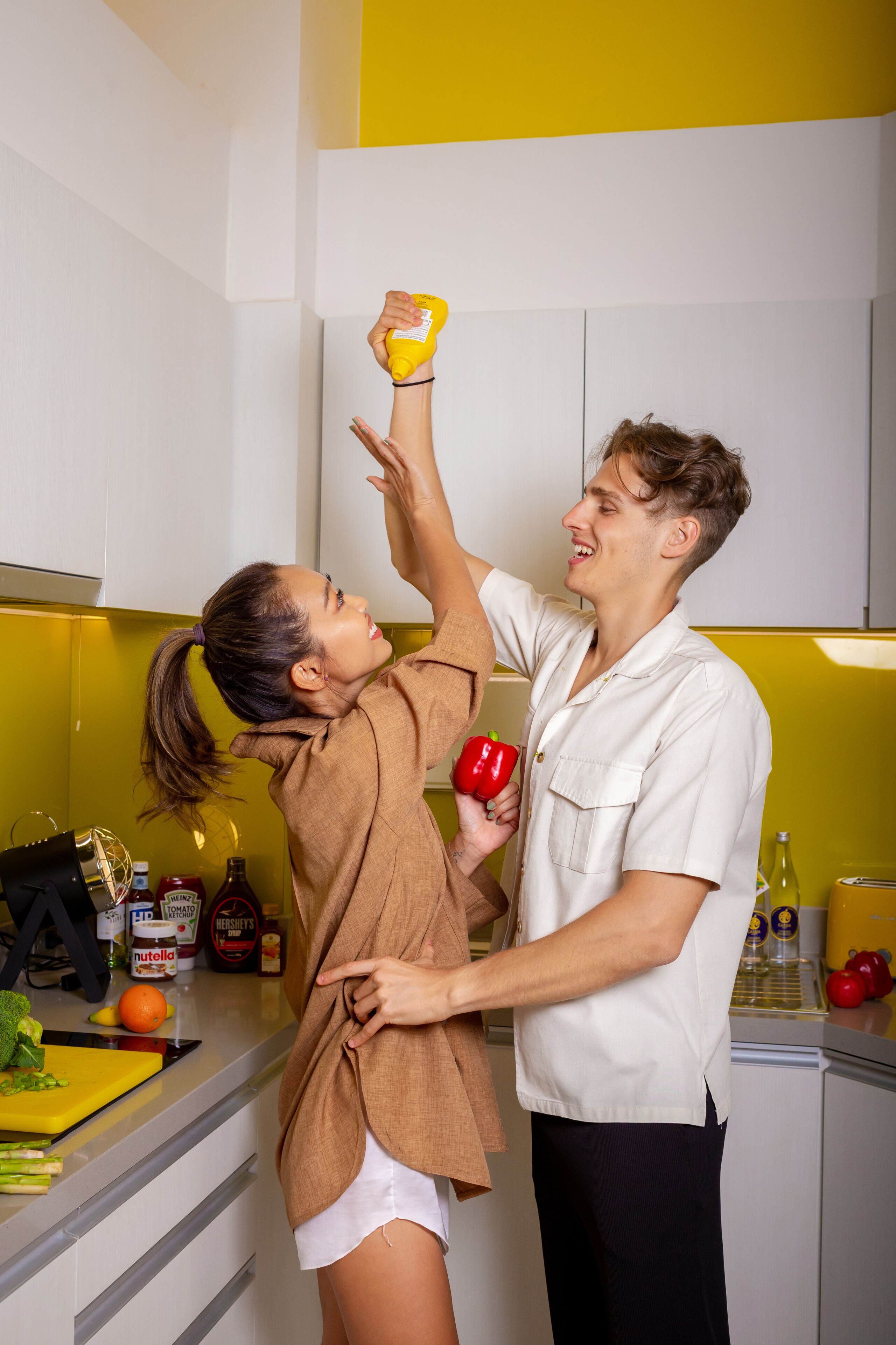 Eine Person hält eine rote Paprika und eine Senfflasche in einer Küche mit weißen Schränken und einer gelben Spritzschutzwand.
