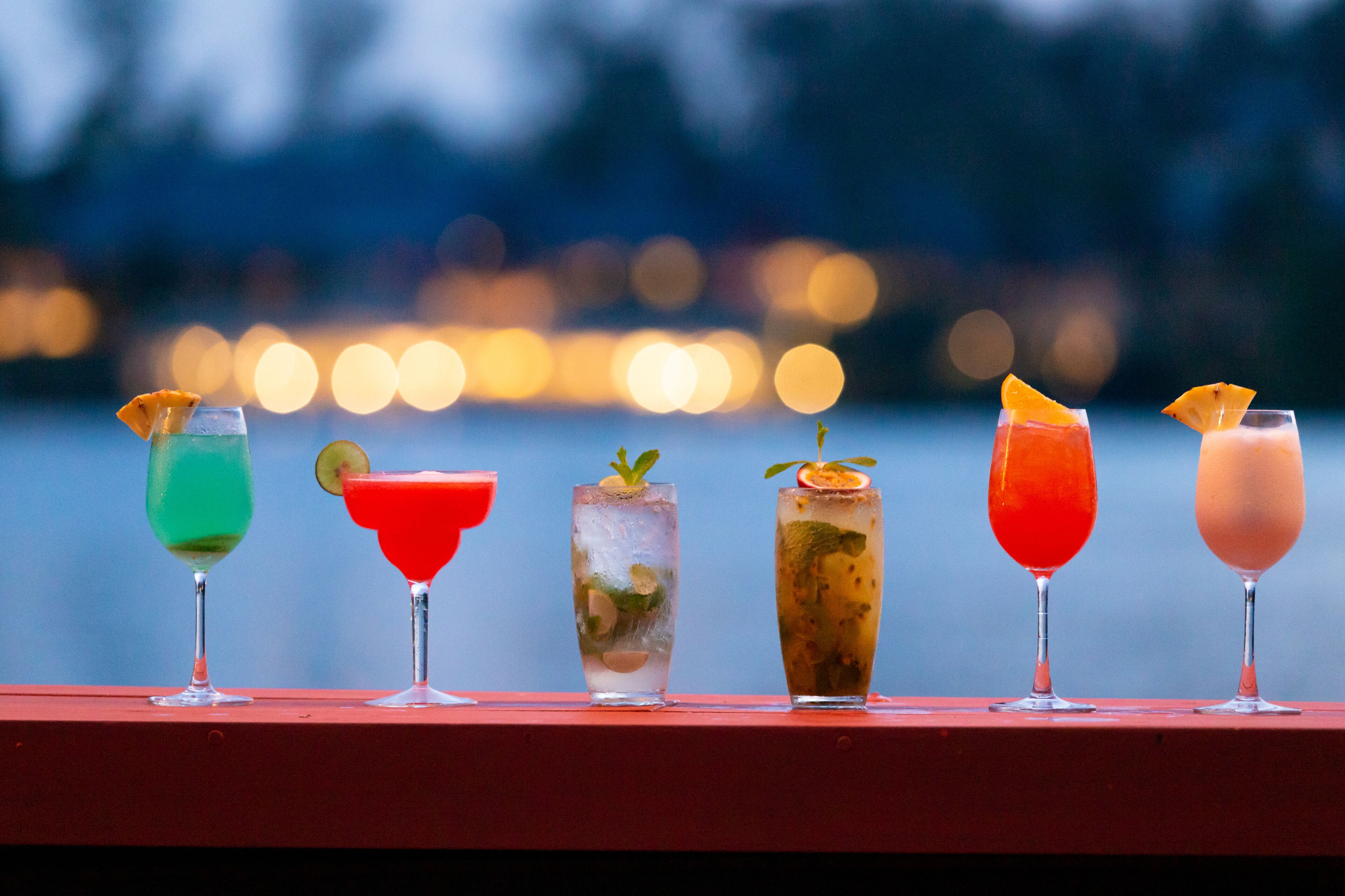 A row of five colorful cocktails on a red surface with a blurred cityscape in the background.