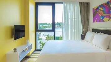 Habitación de hotel con una cama grande, un televisor de pantalla plana montado en la pared, una ventana con vistas a zonas verdes y edificios, y una obra de arte colorida en la pared.