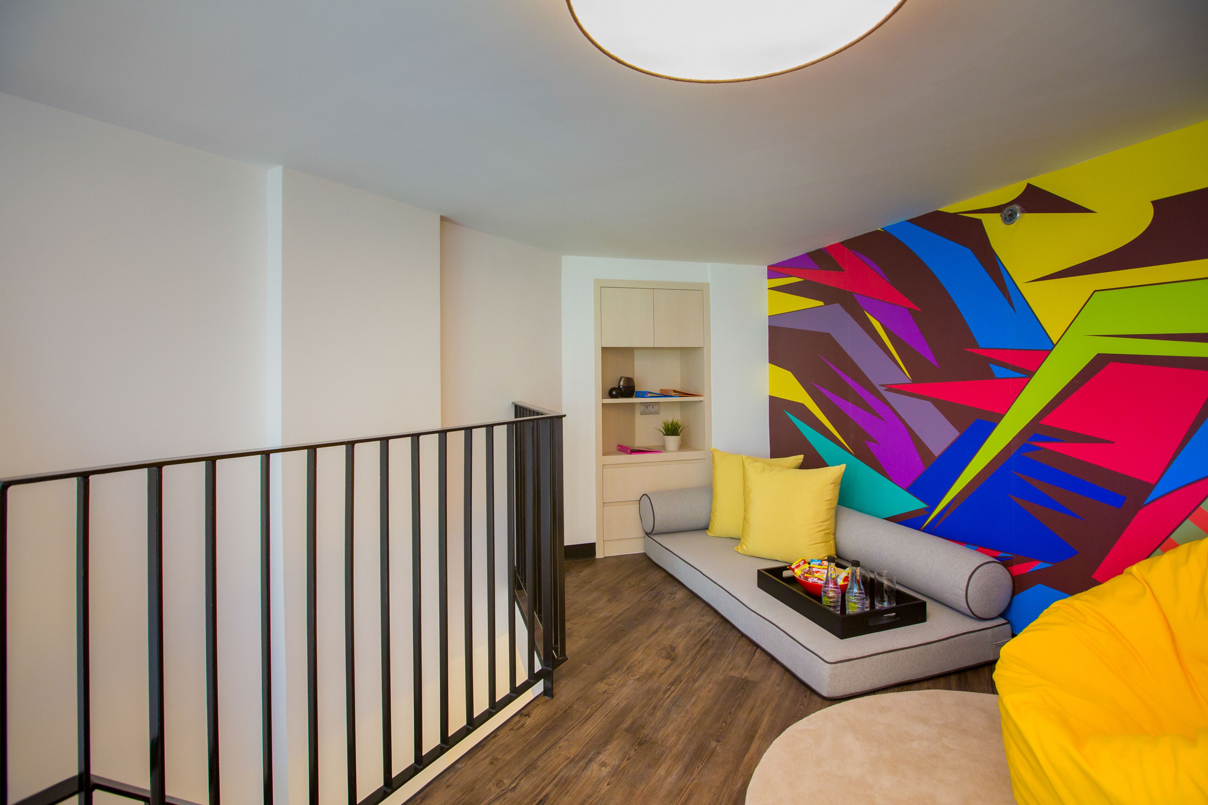 Una sala de estar moderna con suelo de madera, un sofá gris, cojines amarillos y un mural abstracto y colorido en la pared.