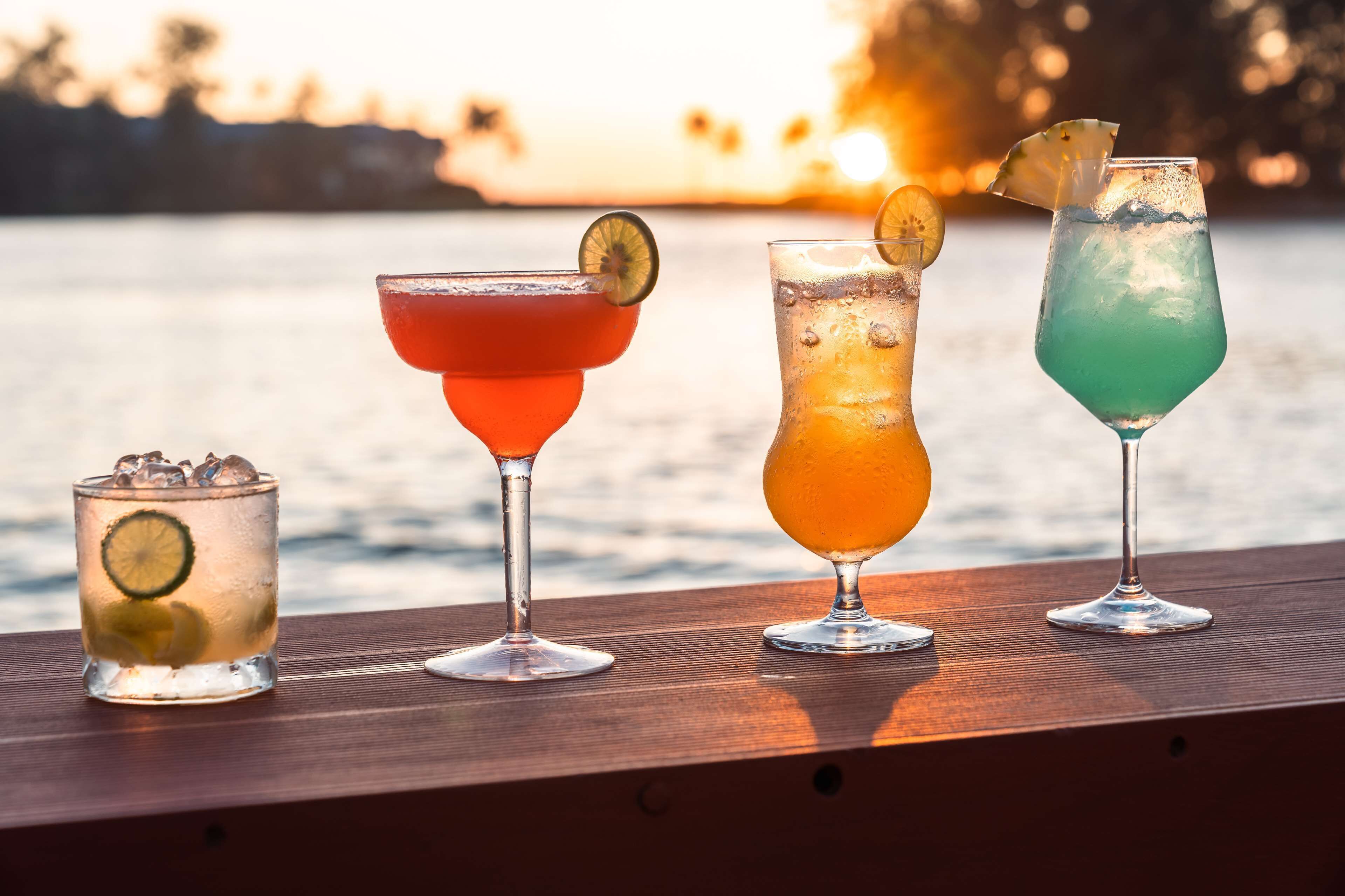 Four cocktails on a wooden table with a body of water in the background.