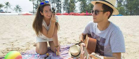 A beach scene with a person playing a guitar and another taking a photo.