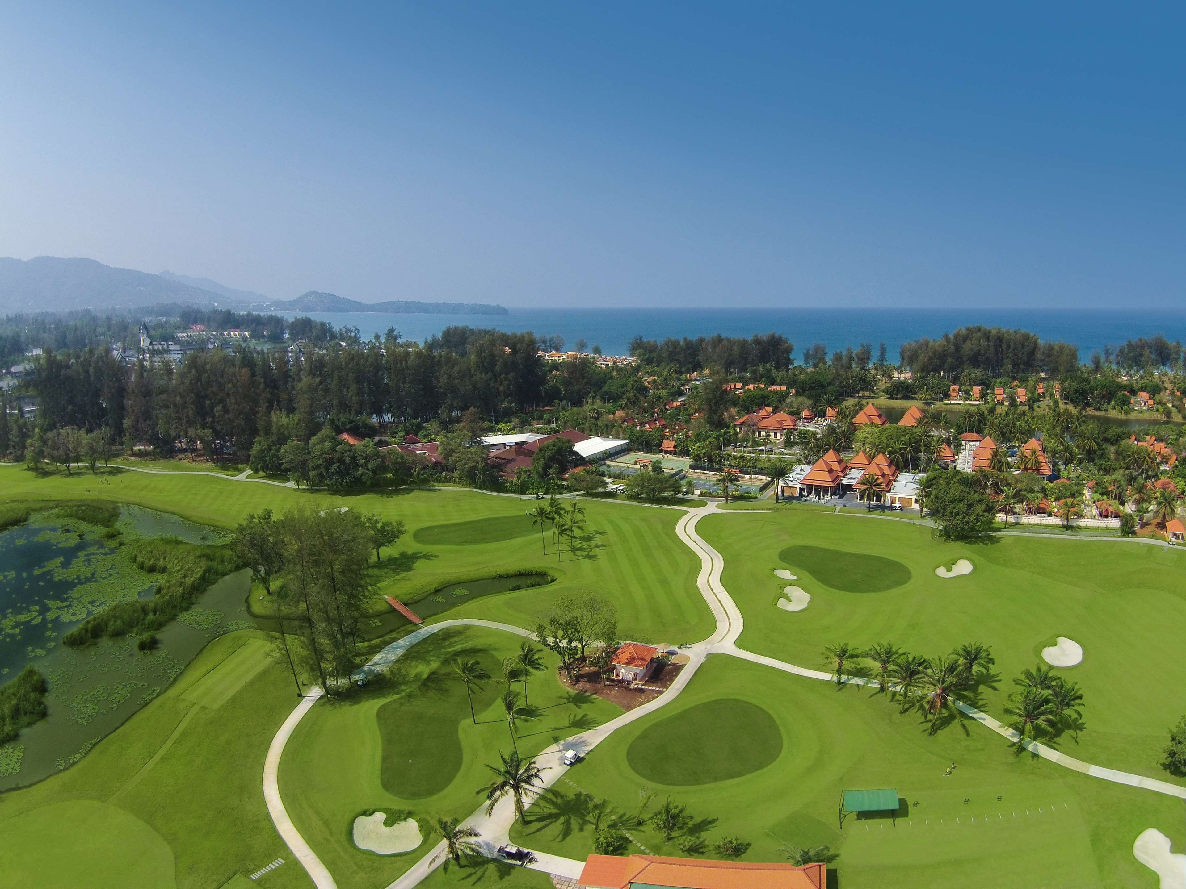 Campo de golf con un cuerpo de agua, una carretera y edificios residenciales.