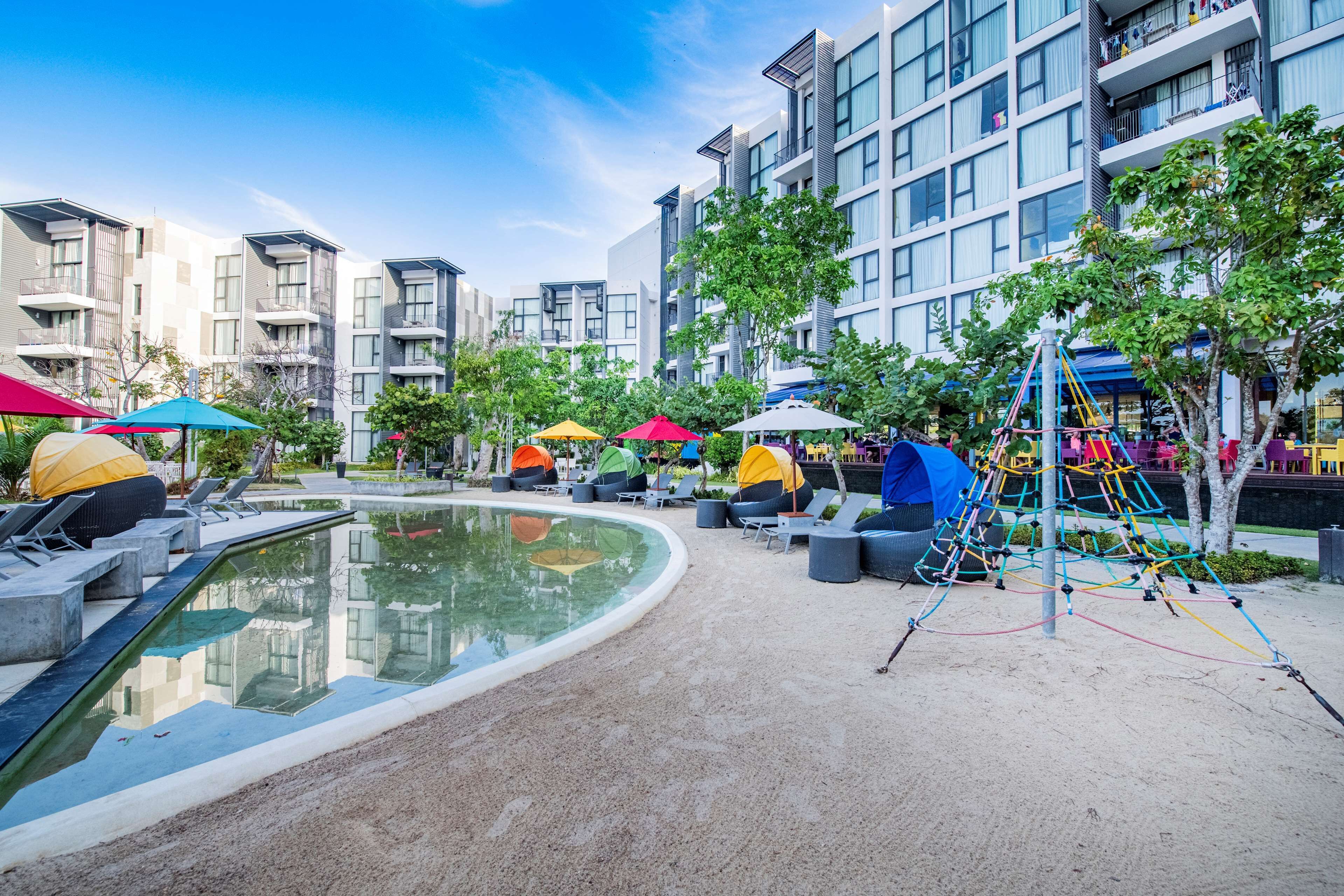 A modern outdoor pool area with lounge chairs and umbrellas, surrounded by multi-story residential buildings.