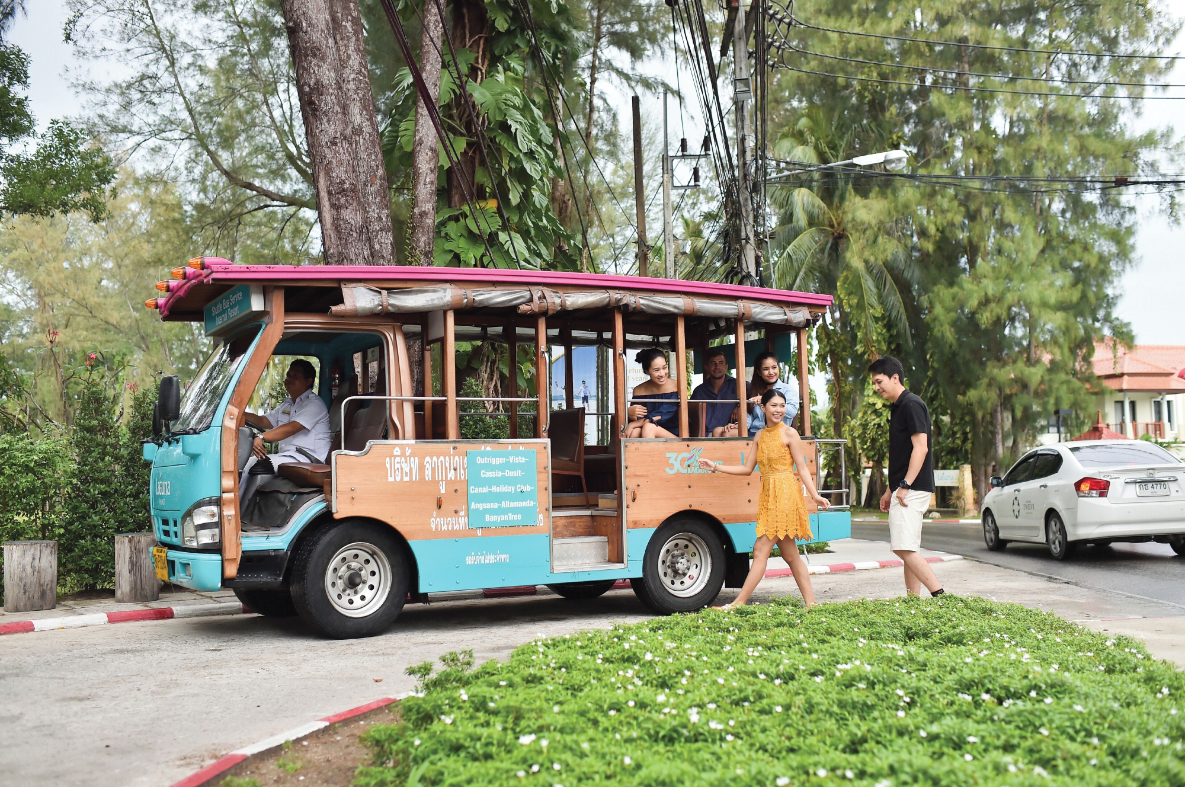 A tuk-tuk with passengers, parked on a street with greenery and a white car in the background.