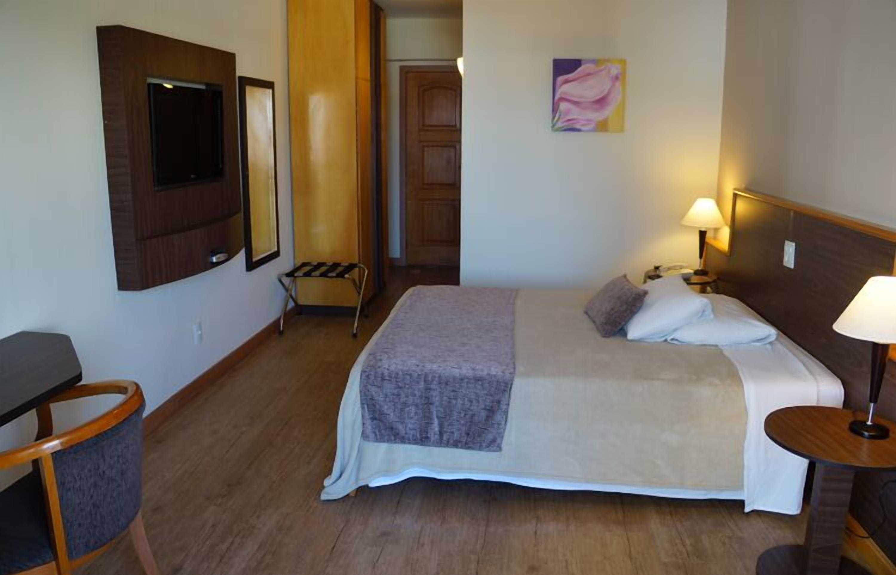 A hotel room with a bed, bedside table, lamp, chair, and a TV mounted on the wall.