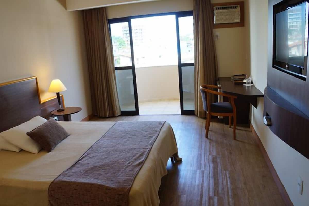 A hotel room with a large bed, a desk, a chair, a TV, and a window with curtains.