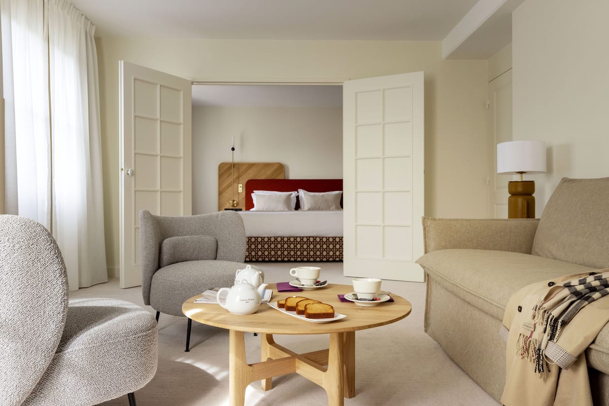 A hotel room with a bed, a sofa, a chair, a small table with a teapot, cups, and bread.