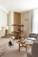 A modern hotel room with a sofa, a round wooden table with tea set, a chair, and a bed with a headboard.