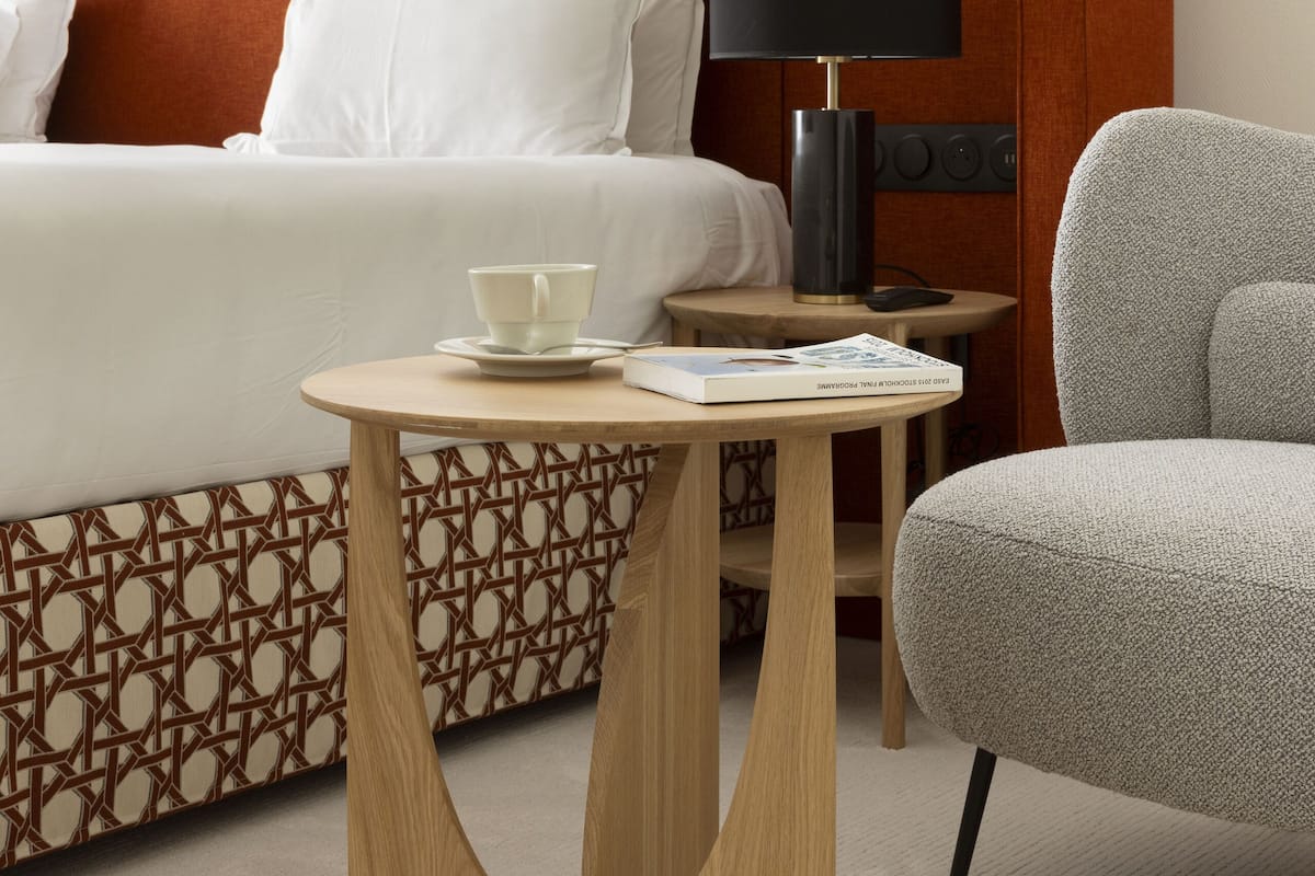 A hotel room with a bed, a bedside table with a lamp, a chair, and a small table with a cup and a magazine.
