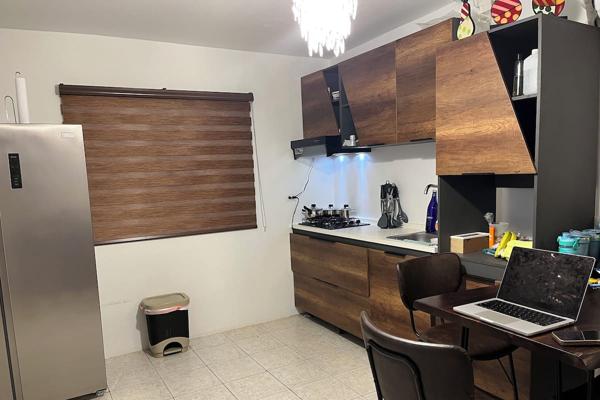 A modern kitchen with a built-in stove, wooden cabinets, a refrigerator, and a laptop on a table.