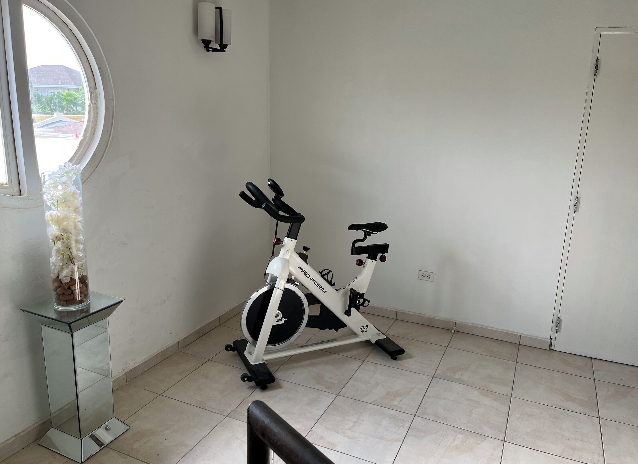 A room with a stationary bike, a glass vase with flowers, and a window with a view of buildings.