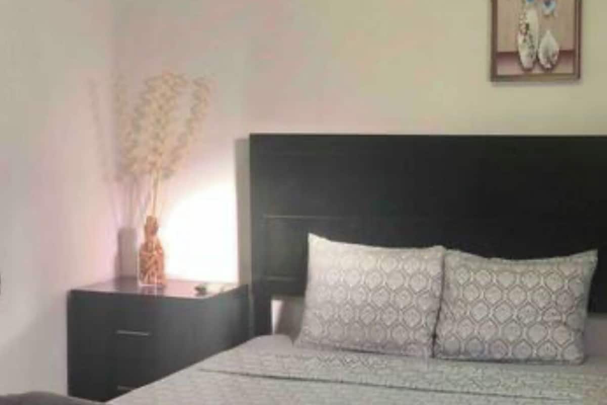 A neatly made bed with patterned pillows, a dark headboard, a bedside table with a vase of flowers, and a framed picture on the wall.