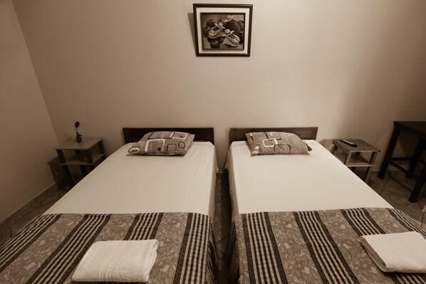 A room with two beds, each with patterned bedding and pillows, a framed artwork on the wall, and a small table with a lamp.