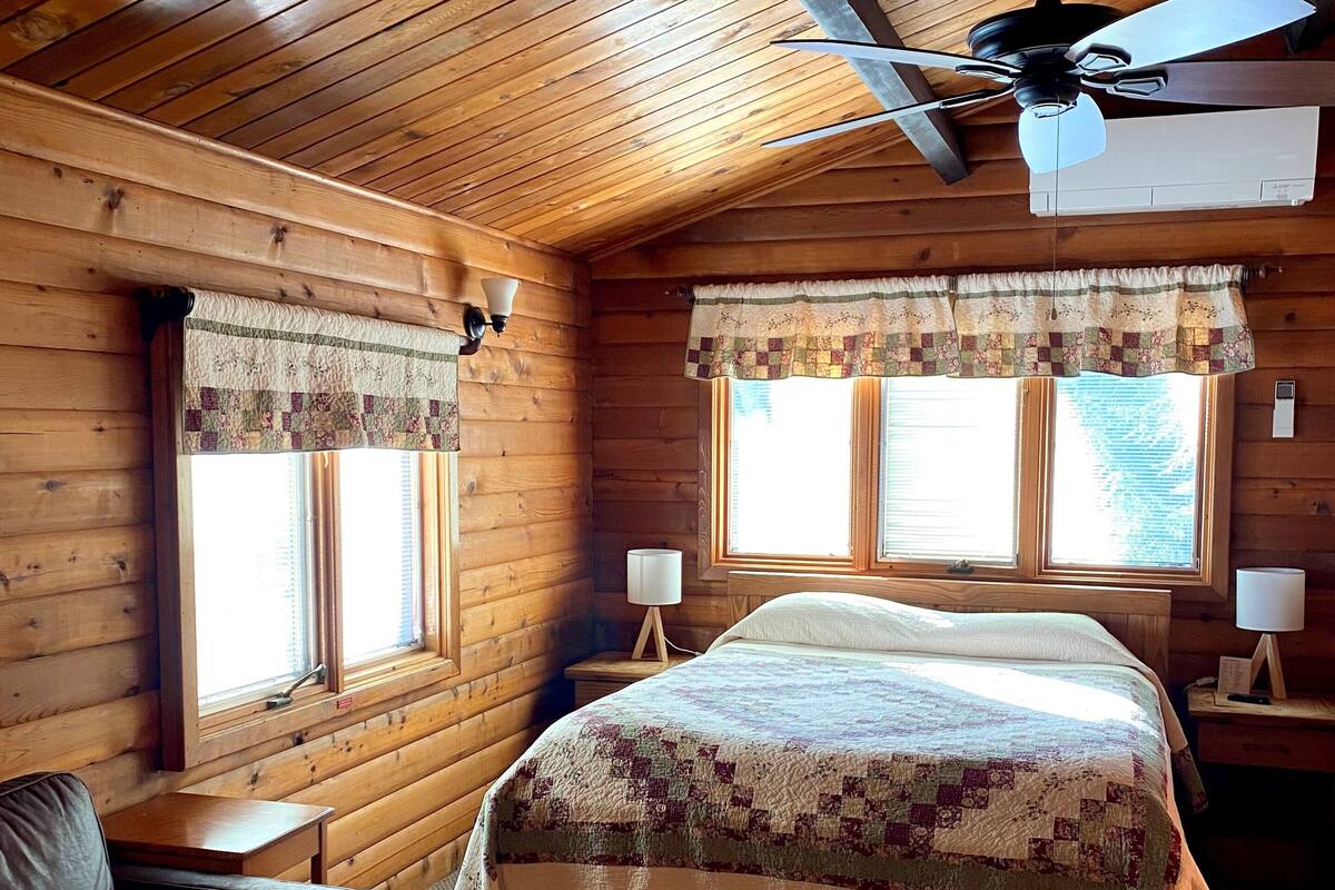 Una habitación acogedora tipo cabaña con techo de madera, una cama con colcha, dos ventanas con cortinas, un ventilador de techo y una mesita de noche con lámpara.