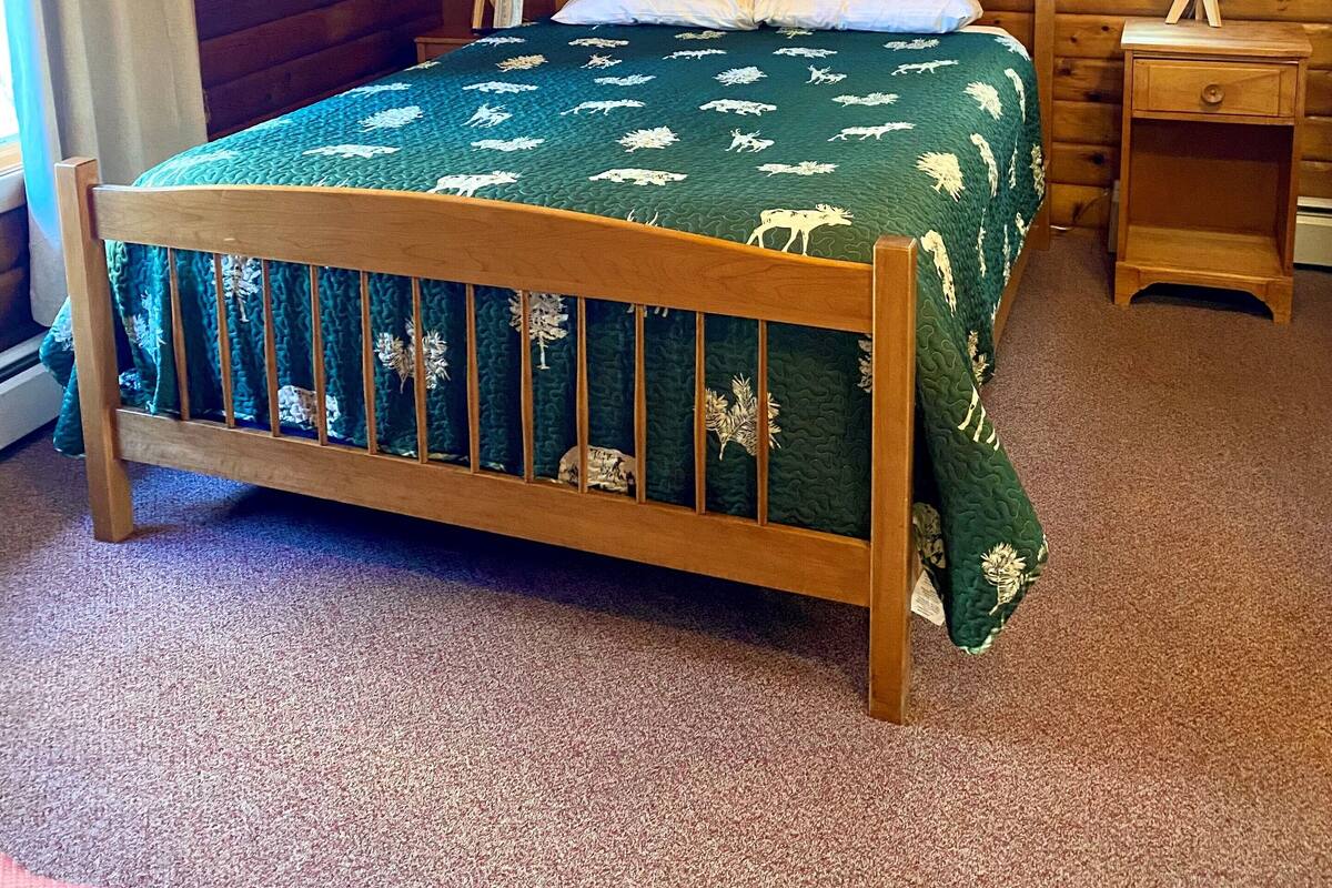 Un dormitorio de cabaña de madera con cama, mesita de noche, ventilador de techo y ventana con cortinas.