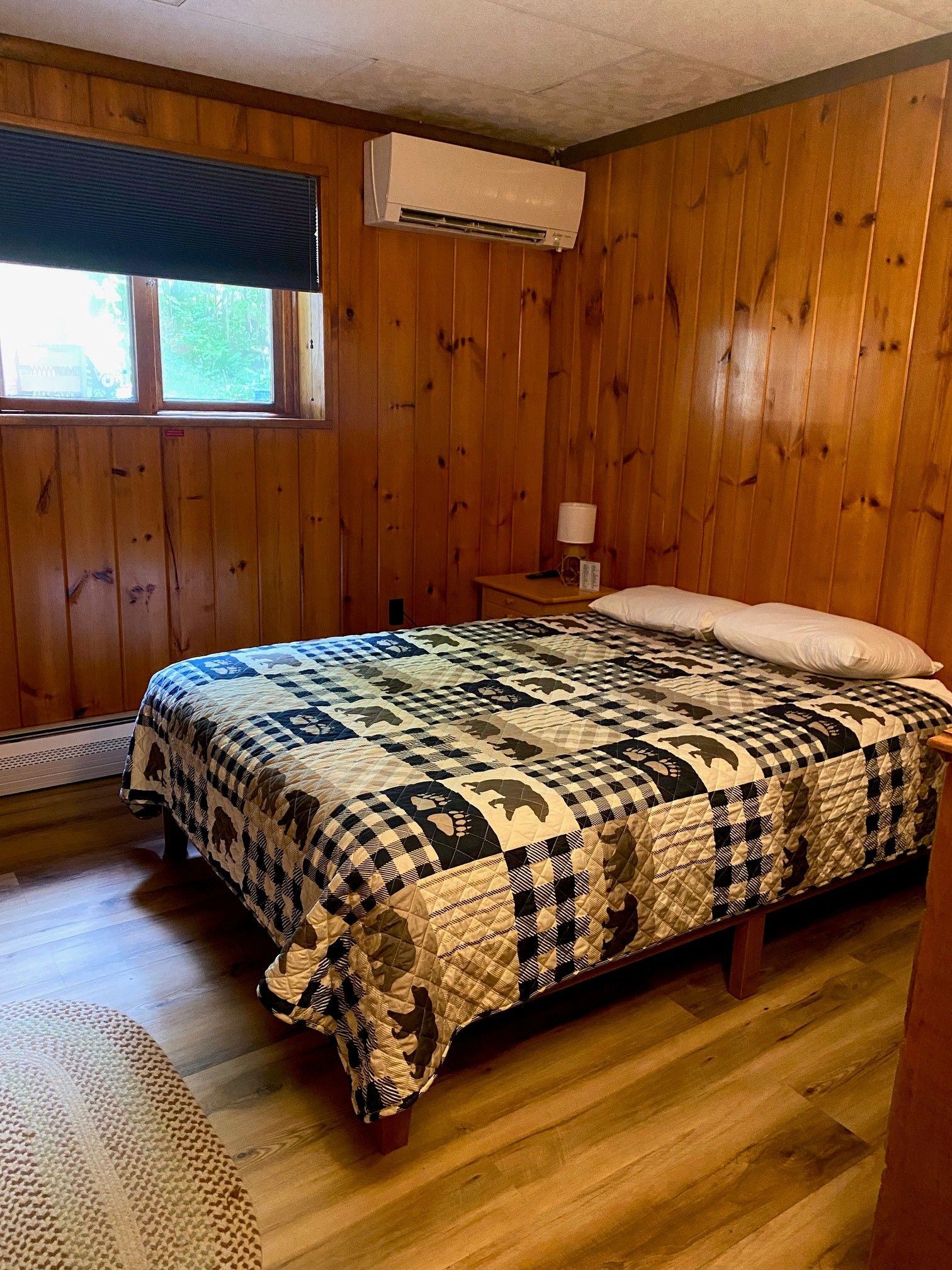 Una habitación con paredes de madera, una cama con colcha, una ventana con persianas y un equipo de aire acondicionado.