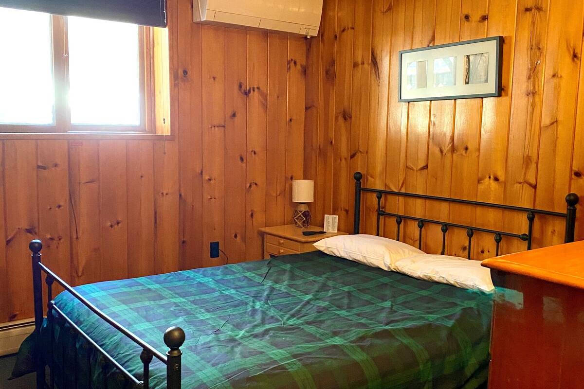 Una habitación con paredes de madera, una cama con colcha verde y negra a cuadros, una mesita de noche con lámpara y un cuadro en la pared.