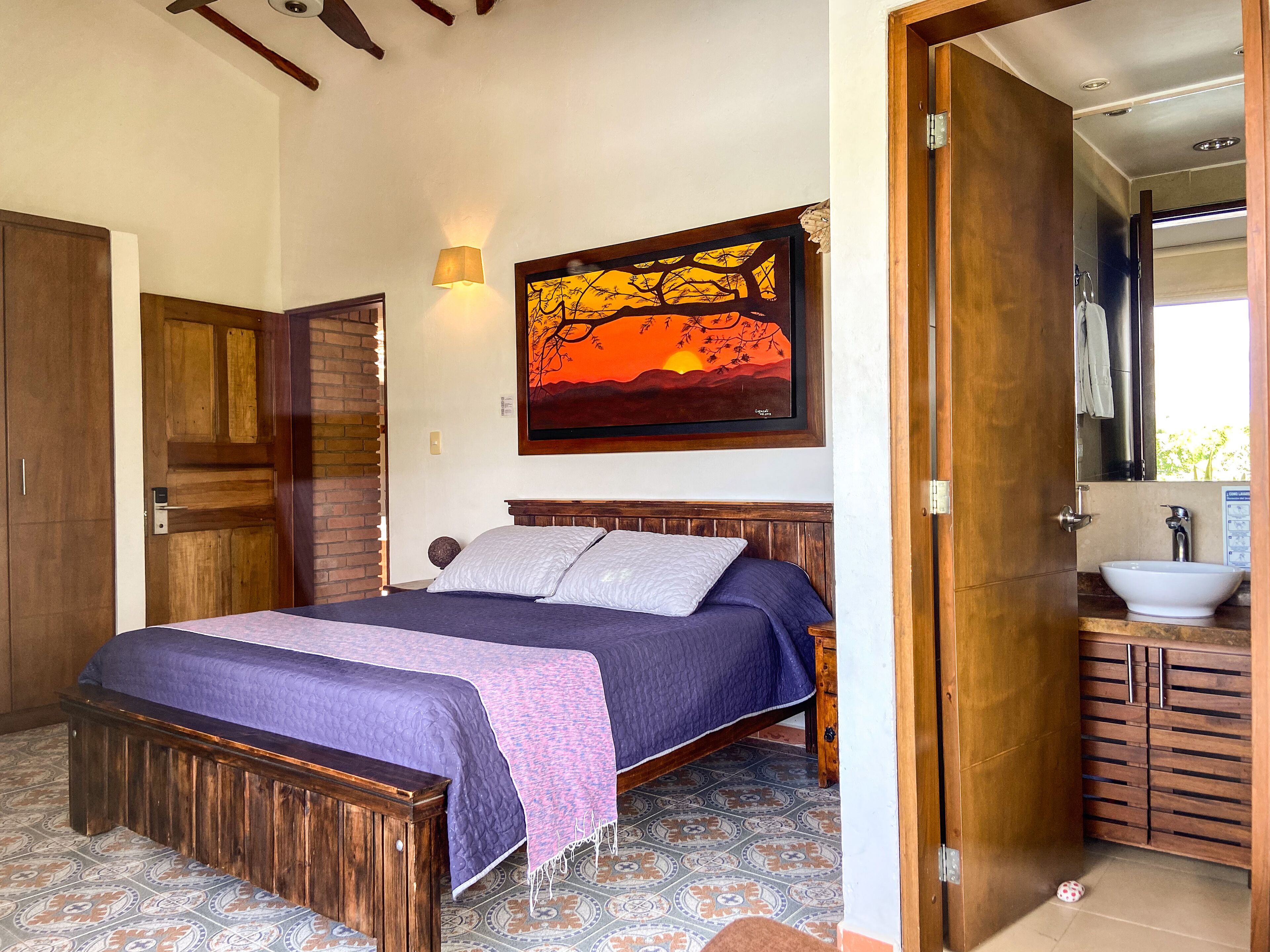 Une chambre à coucher avec un lit en bois, une table de chevet, un tableau accroché au mur, et une salle de bain avec un lavabo et un miroir.