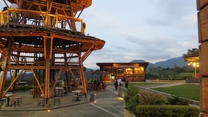 Una estructura de bambú con asientos exteriores, un edificio de cristal con techo rojo y vistas a montañas al fondo.