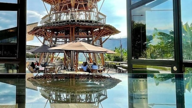 Un área de estar exterior moderna con estructura de madera y sombrillas, reflejada en un suelo de vidrio.