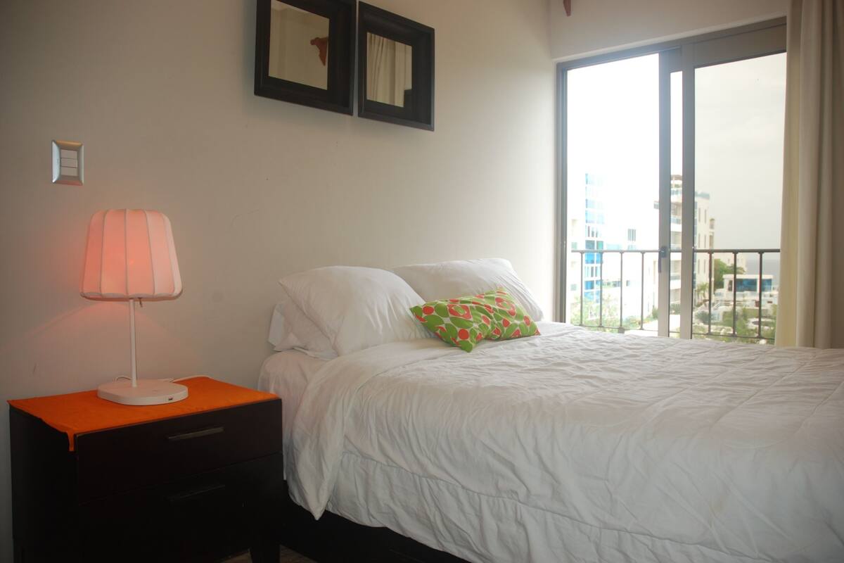 A bedroom with a bed, a nightstand, a lamp, and a window with a view of buildings.