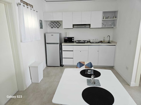 Cocina funcional con armarios blancos, refrigerador, microondas y lavamanos. Se aprecia una pequeña mesa de comedor con sillas.