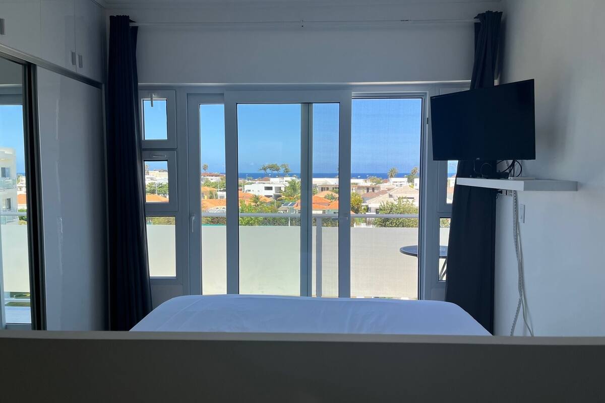 A hotel room with a bed, a television on a stand, and a balcony with a view of buildings and the sea.