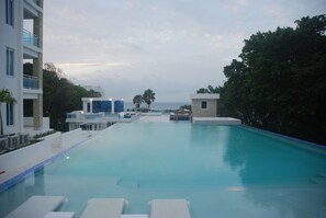 A swimming pool with lounge chairs, a view of the sea, and modern buildings. - E&J Beach Condos (Sosúa)