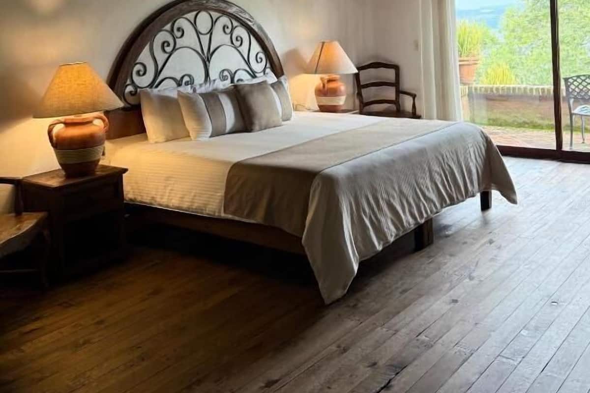Une chambre à coucher avec un plafond en bois, un lit doté d’une tête de lit, deux tables de chevet avec des lampes, et une vue dégagée sur l’extérieur grâce à une grande fenêtre.