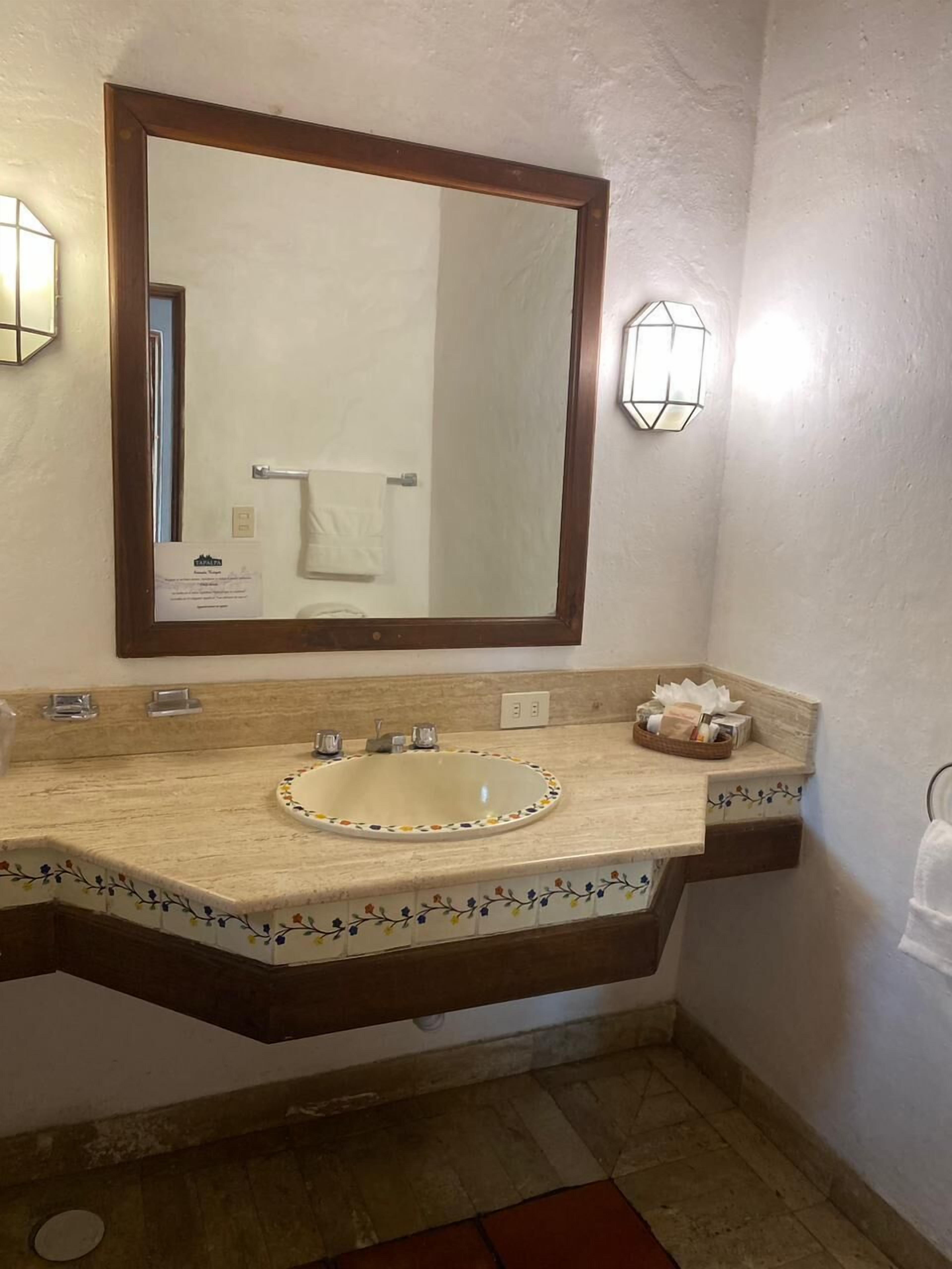 A bathroom with a large mirror, a sink with a decorative border, a towel rack, and a wall-mounted light fixture.