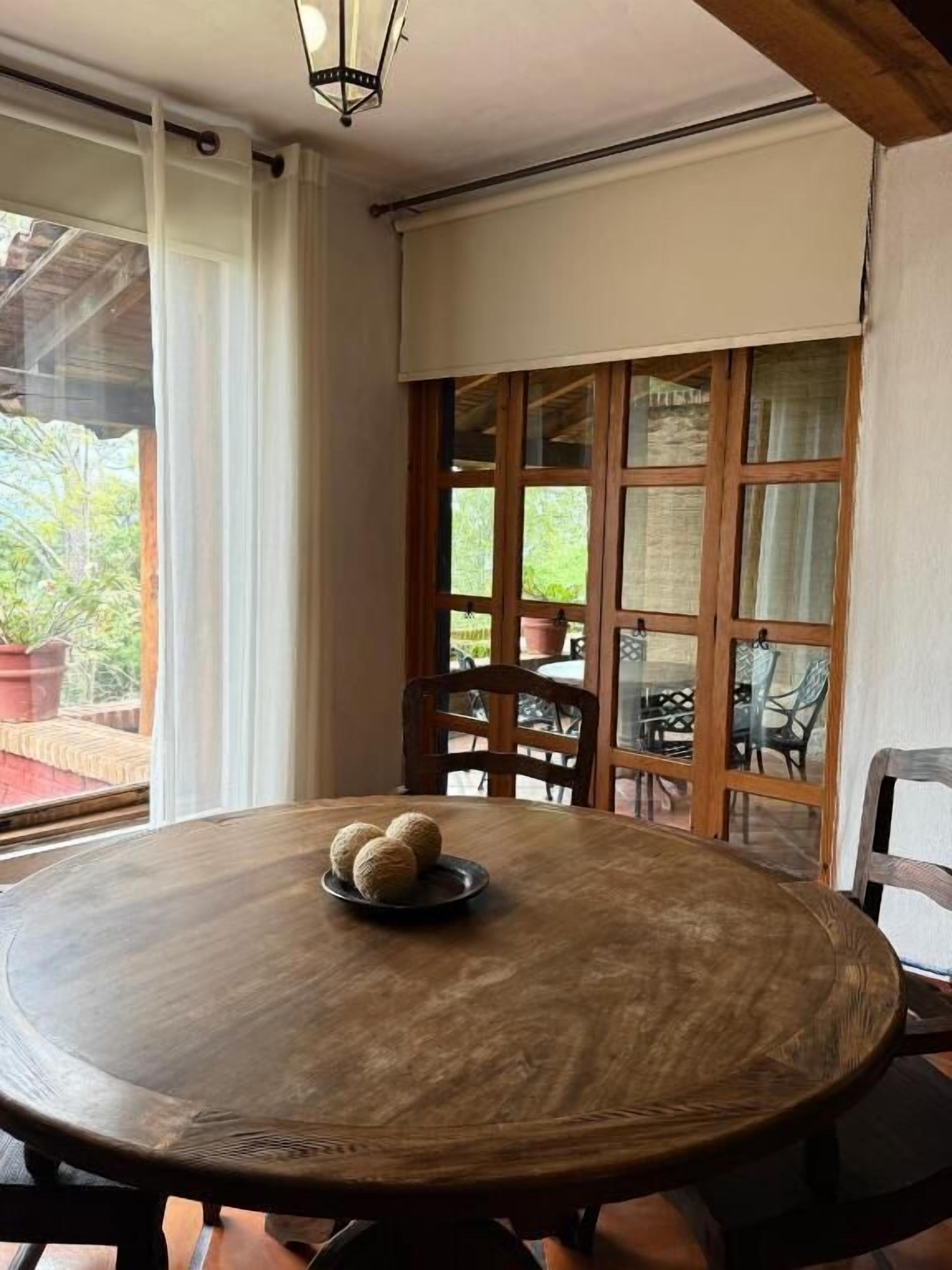 A round wooden table with a plate of three round objects, a wooden chair, and a view of an outdoor area with chairs and potted plants.
