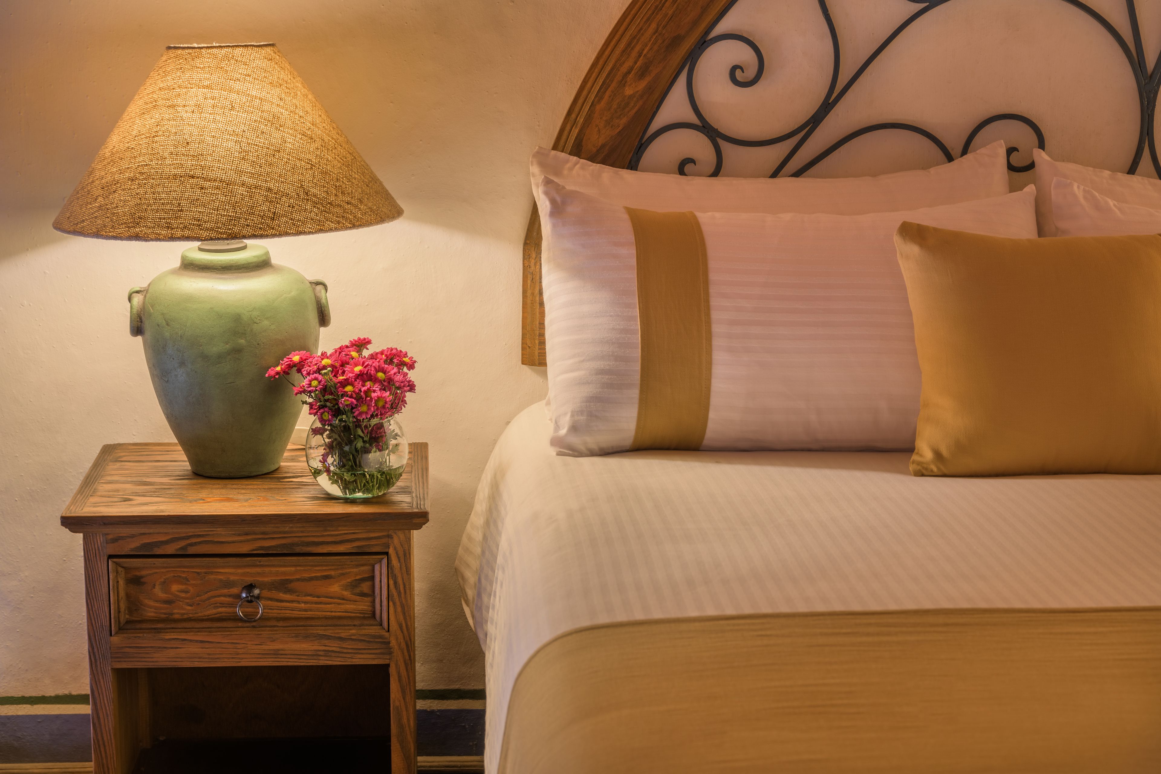A neatly made bed with a headboard, a bedside table with a lamp, and a vase with flowers.