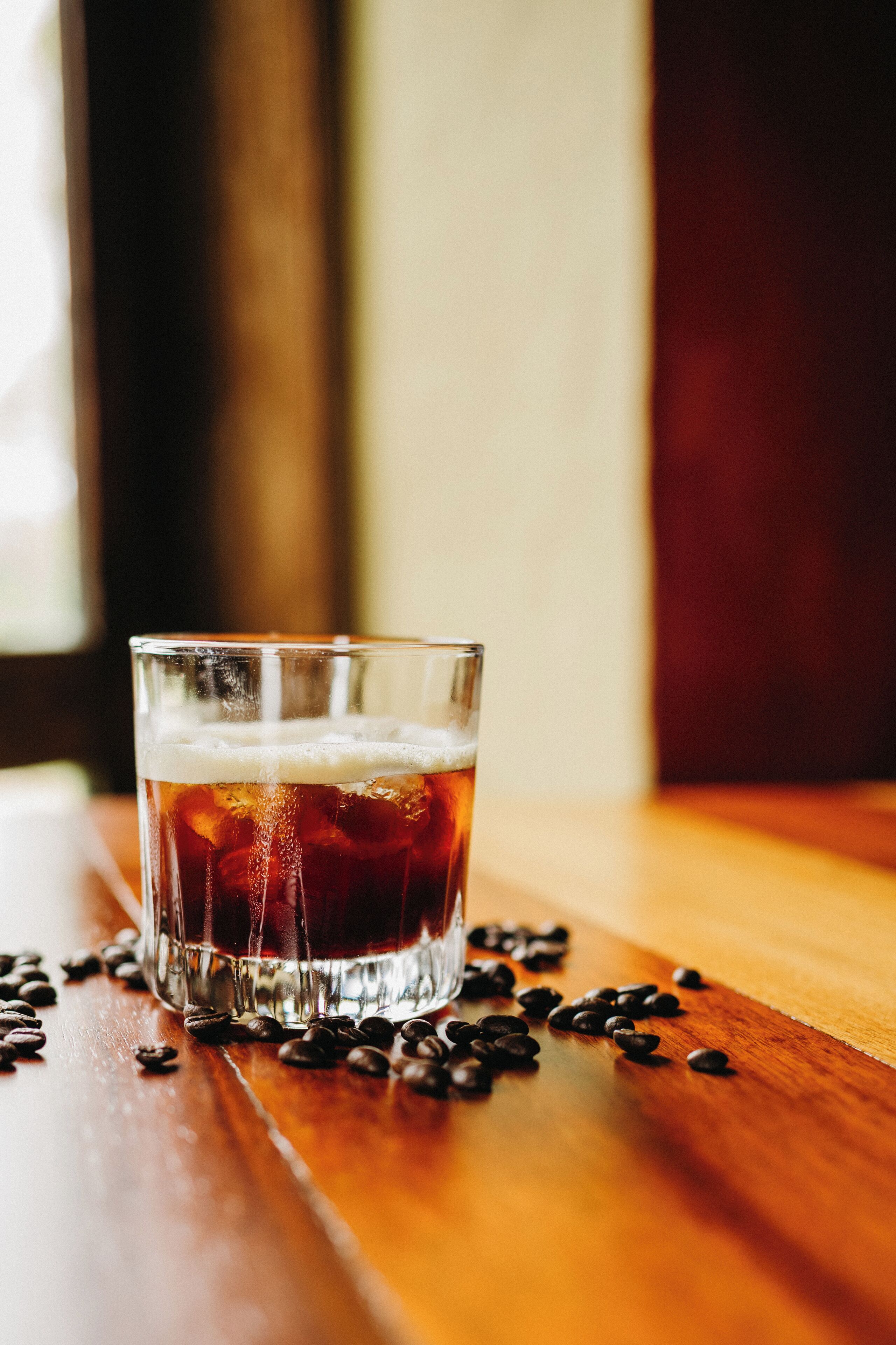 A glass of iced tea with coffee beans scattered around it on a wooden surface.