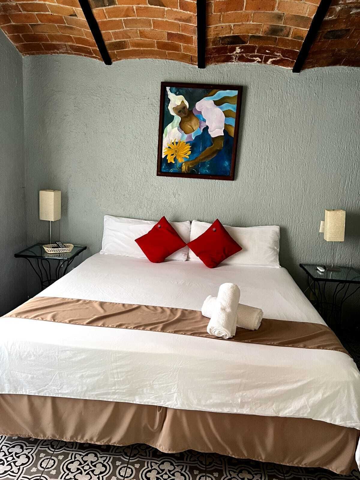 Habitación de hotel con cama, dos almohadas rojas, una toalla blanca, un cuadro en la pared y una alfombra estampada.