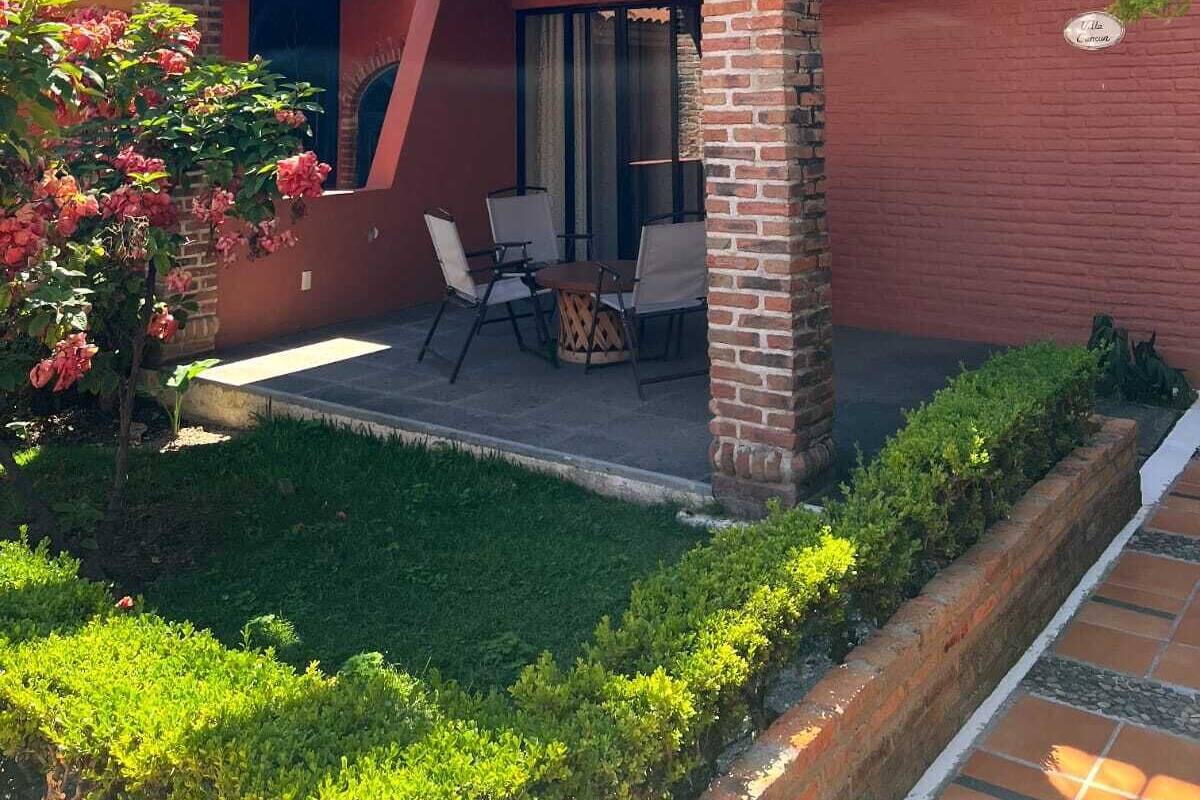 Un edificio de dos pisos con un balcón, un patio con sillas y una zona ajardinada.