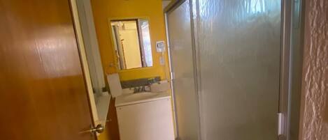 A narrow hallway with a white refrigerator, a mirror, and a door leading to a room.