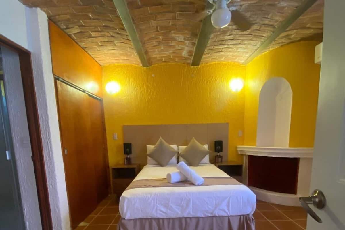 Habitación de hotel con techo de ladrillo, cama con ropa blanca, mesita de noche con lámpara y chimenea empotrada en la pared.