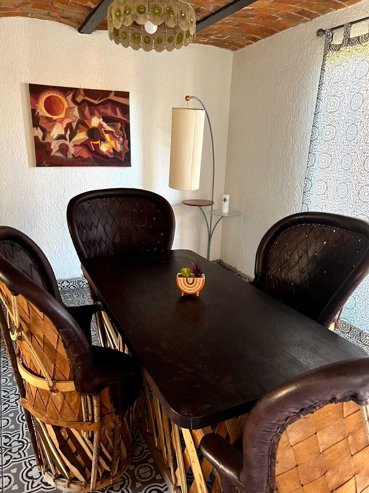 A dining area with a dark wooden table, four cushioned chairs, a small decorative bowl, a wall-mounted lamp, and a painting.