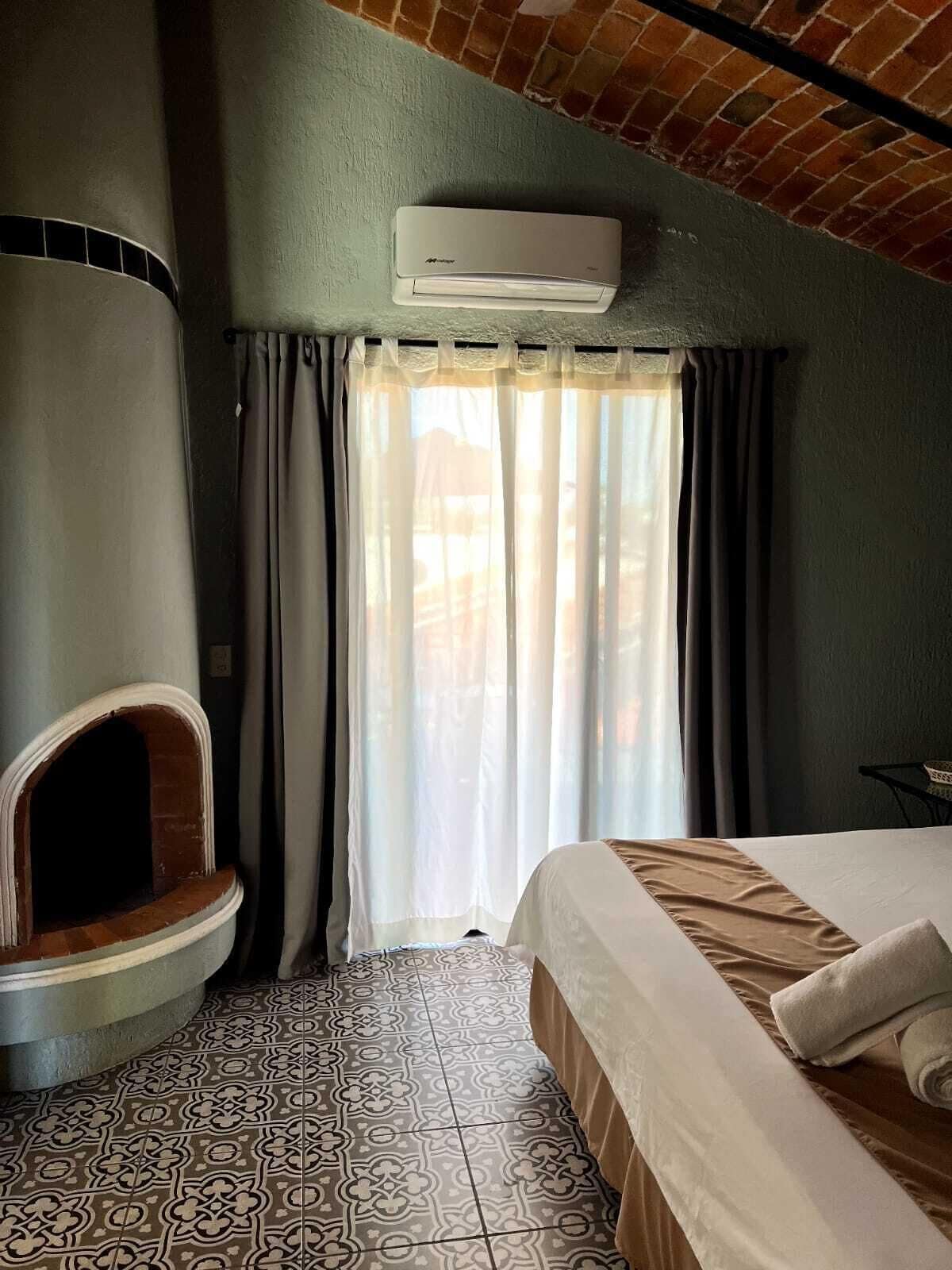 A room with a bed, a built-in fireplace, patterned floor tiles, and a window with curtains.