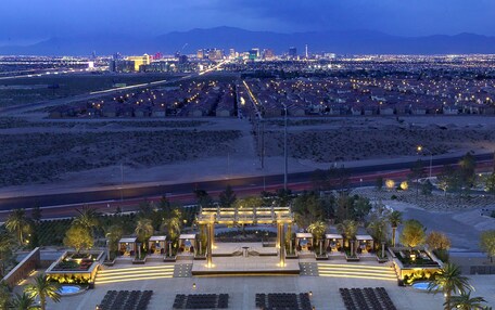 A large outdoor swimming pool with multiple lounge areas and seating arrangements, surrounded by palm trees and a view of a cityscape in the background.