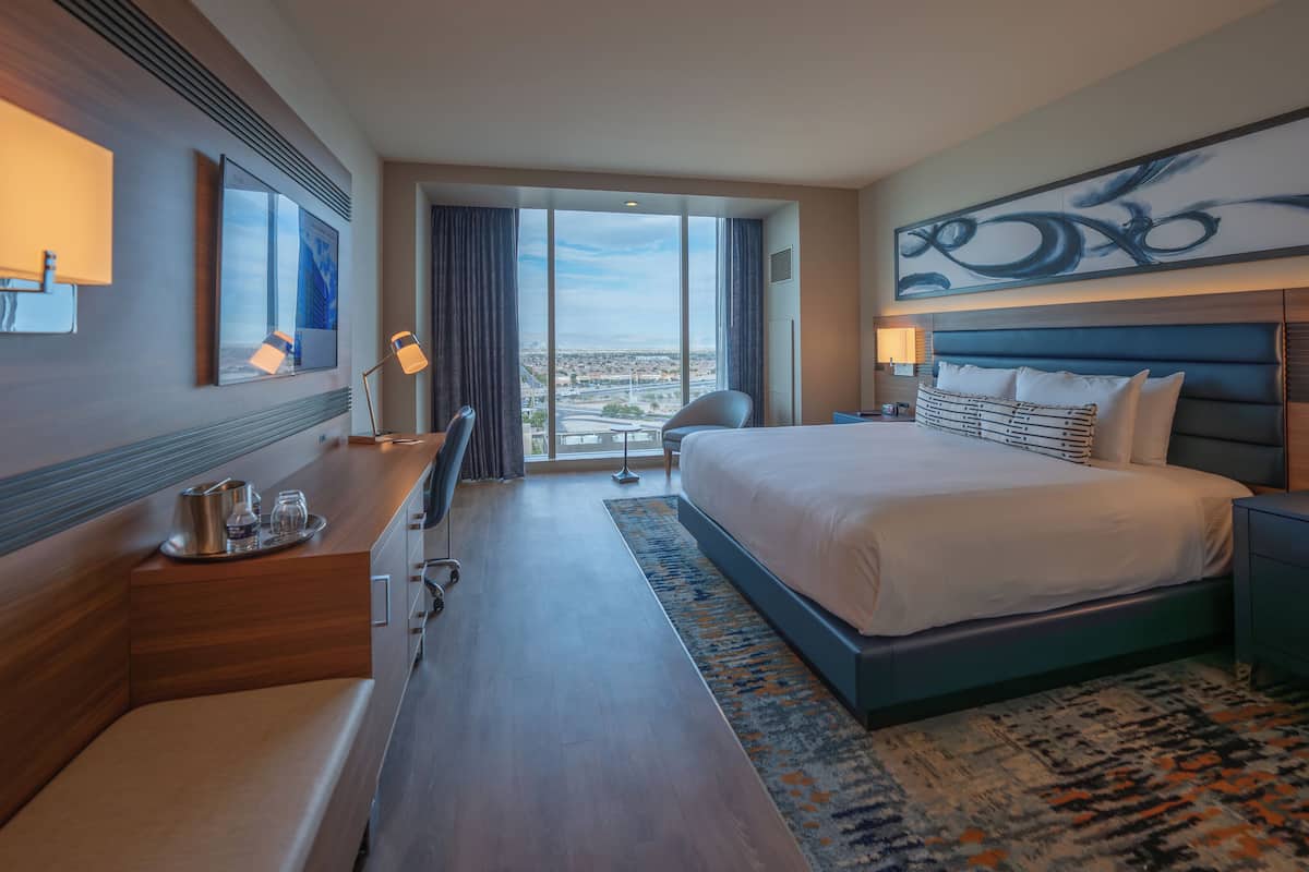 Een moderne hotelkamer met een groot bed, een bureau met een lamp, een stoel en stadszicht door grote ramen.