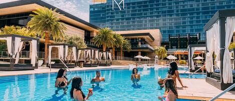 A swimming pool area with lounge chairs and umbrellas, surrounded by palm trees and a modern building with a glass facade.