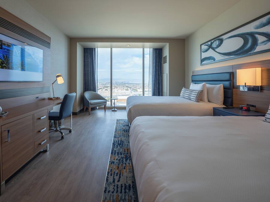 A hotel room with a large bed, a desk with a chair, a TV on the wall, and a view of the city through the windows.