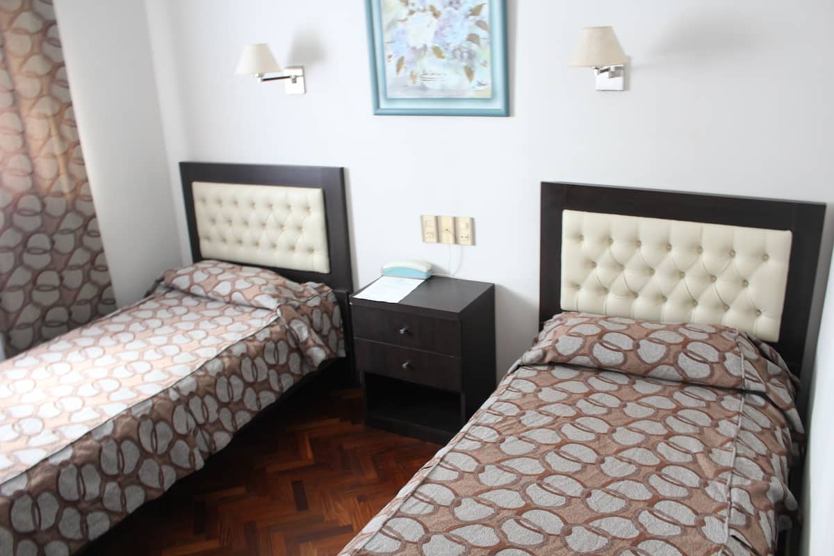 Habitación de hotel con dos camas individuales, una mesita de noche, una lámpara y un cuadro en la pared.