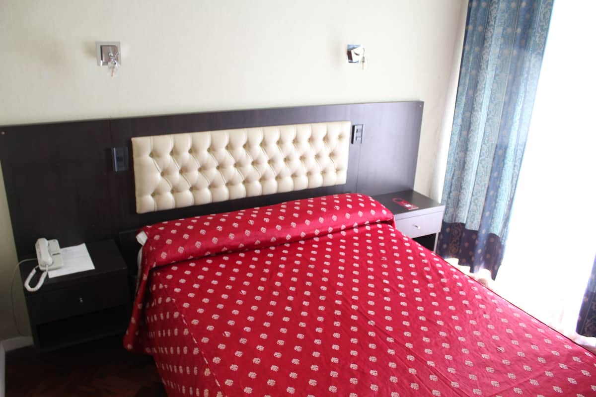 Una habitación de hotel con un cabecero capitonado, una colcha roja con un diseño blanco, una mesita de noche con un teléfono blanco y otra mesita con un teléfono blanco y un papel.