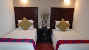 Une chambre d’hôtel avec deux lits, chacun recouvert d’une courtepointe blanche et d’une couverture colorée. Il y a deux tables de chevet avec des lampes, et une petite horloge sur l’une d’elles.