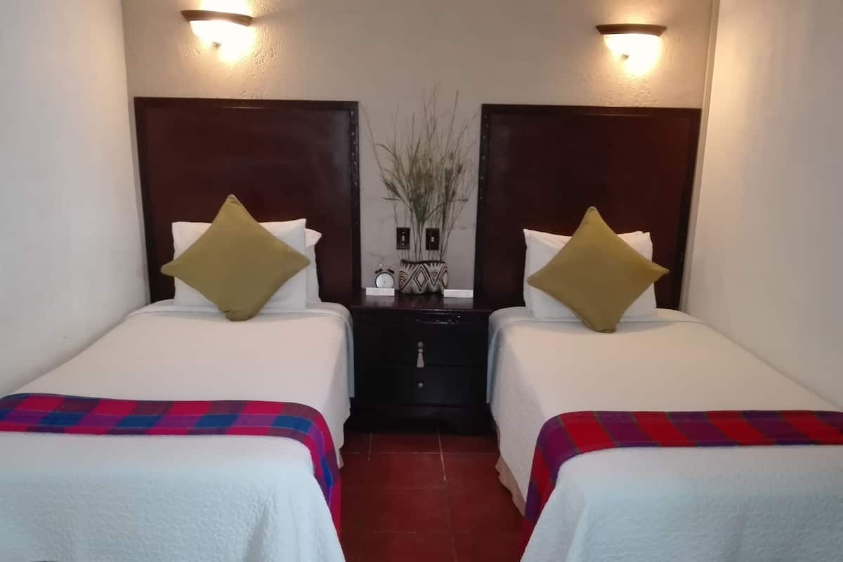 Une chambre d’hôtel avec deux lits, chacun recouvert d’une courtepointe blanche et d’une couverture colorée. Il y a deux tables de chevet avec des lampes, et une petite horloge sur l’une d’elles.