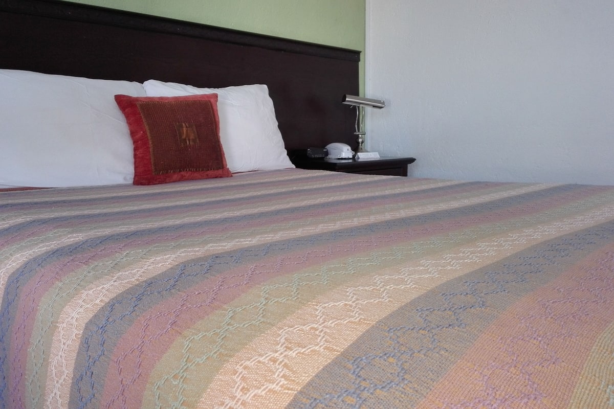 Un lit bien fait, recouvert d’une couvre-lit à rayures, avec un oreiller rouge et une table de chevet sur laquelle se trouvent une lampe et un téléphone.